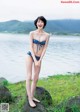 A woman in a blue bikini standing on a rock by the water.